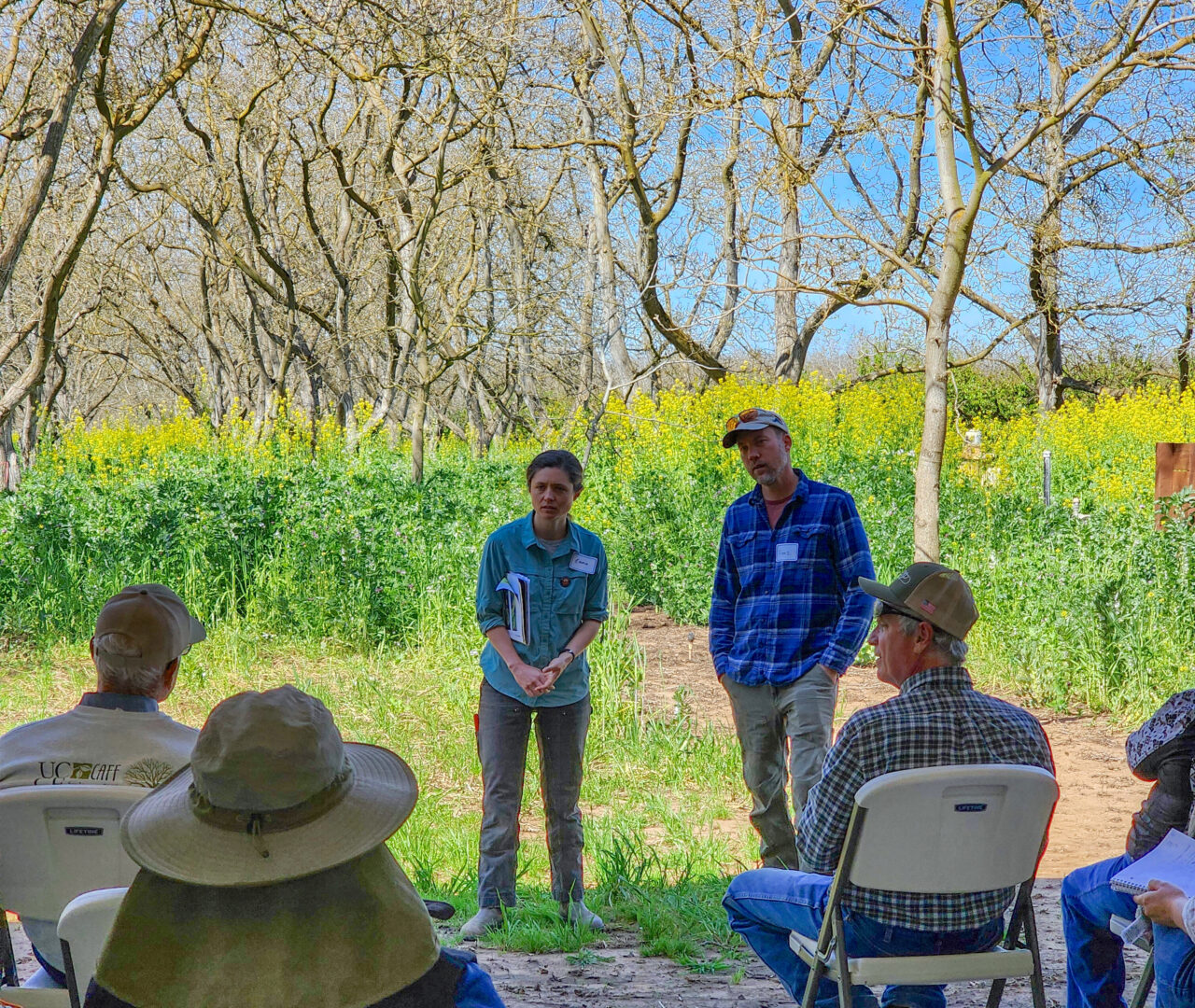 Farmers Franz Eilers and Emma Wade standing in front of attendees, with a tall, blooming cover crop and walnut trees in the background.